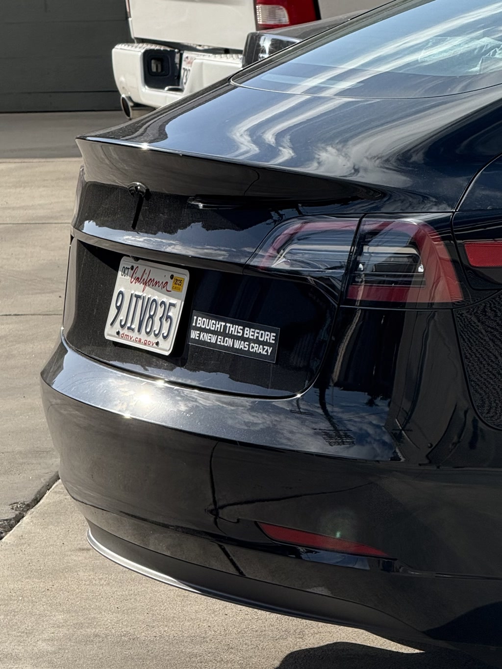 A sleek black car with a glossy finish is parked outdoors. The car's rear end is prominently visible, showcasing its distinctive taillights. A California license plate is attached to the car, displaying the registration number. Additionally, there is a sticker on the bumper with the text, 'I bought this before we knew Elon was crazy.'