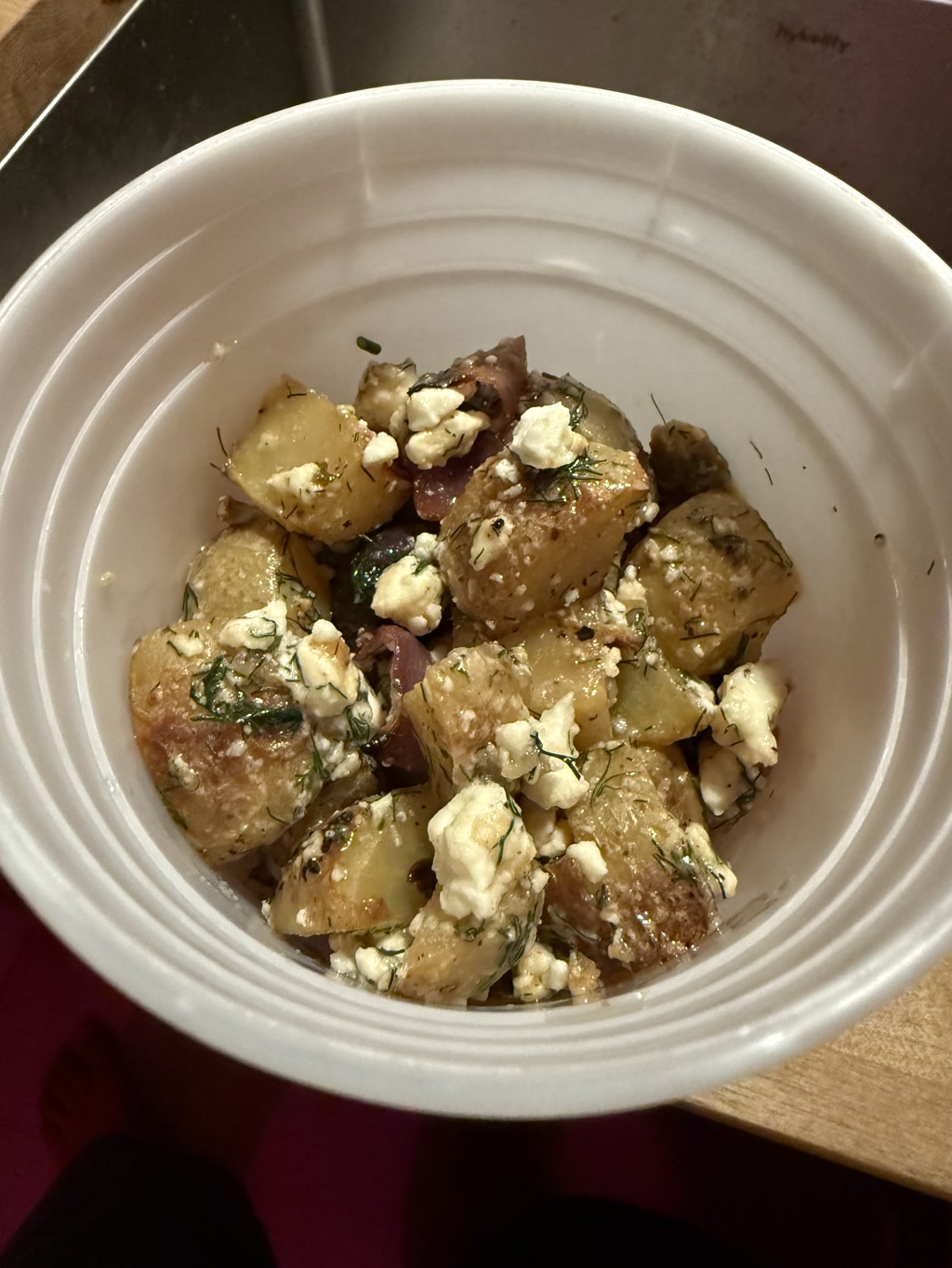 A white bowl filled with diced potatoes that are generously topped with crumbled white cheese and finely chopped herbs. The bowl is placed on a wooden surface, and there is a knife visible in the background. The potatoes appear to be well-seasoned, with a mix of larger chunks and smaller pieces, creating a visually appealing and appetizing dish.