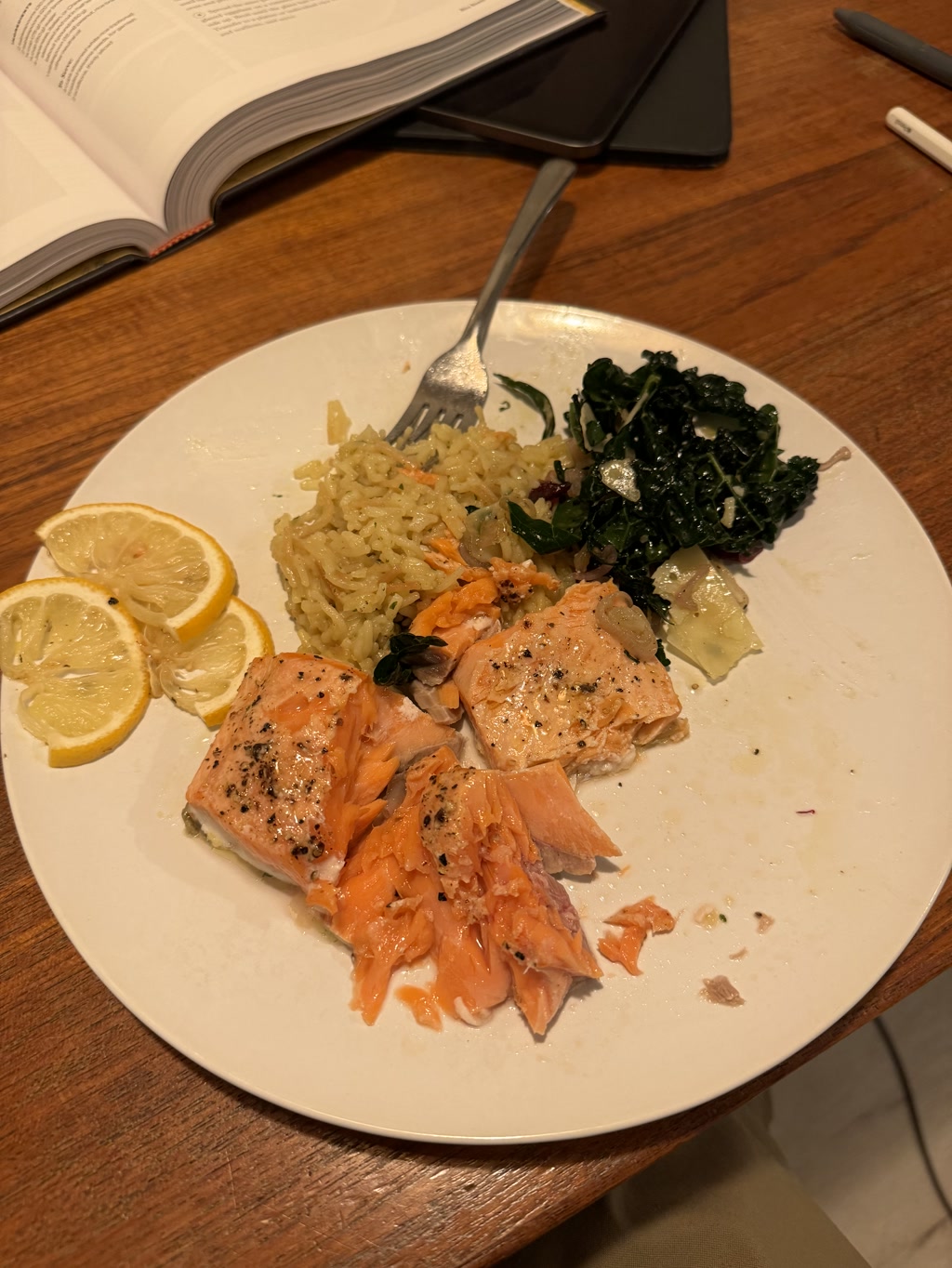 A plate of food is partially eaten. There are a few pieces of flaky cooked salmon seasoned with pepper, a side of rice pilaf, and a serving of dark leafy greens which could be spinach or kale. Two wedges of lemon are also on the plate, presumably for adding to the salmon or the greens. In the background, an open book, a closed notebook and a writing implement rest on a wooden table, suggesting a study or work environment where a meal is being enjoyed.