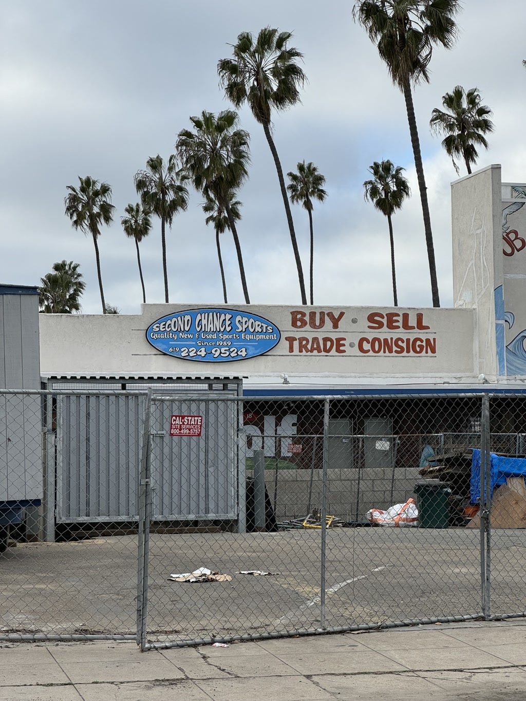 The photo shows the façade of a building with a large sign reading 'SECOND CHANCE SPORTS,' indicating a business that deals in quality new and used sports equipment since 1999. The sign also prominently features the options to 'BUY SELL TRADE CONSIGN.' A chain-link fence surrounds the area, with various objects and debris visible on the ground. In the background, against a cloudy sky, a row of tall palm trees stands above the building, contributing to a distinctly Californian atmosphere.