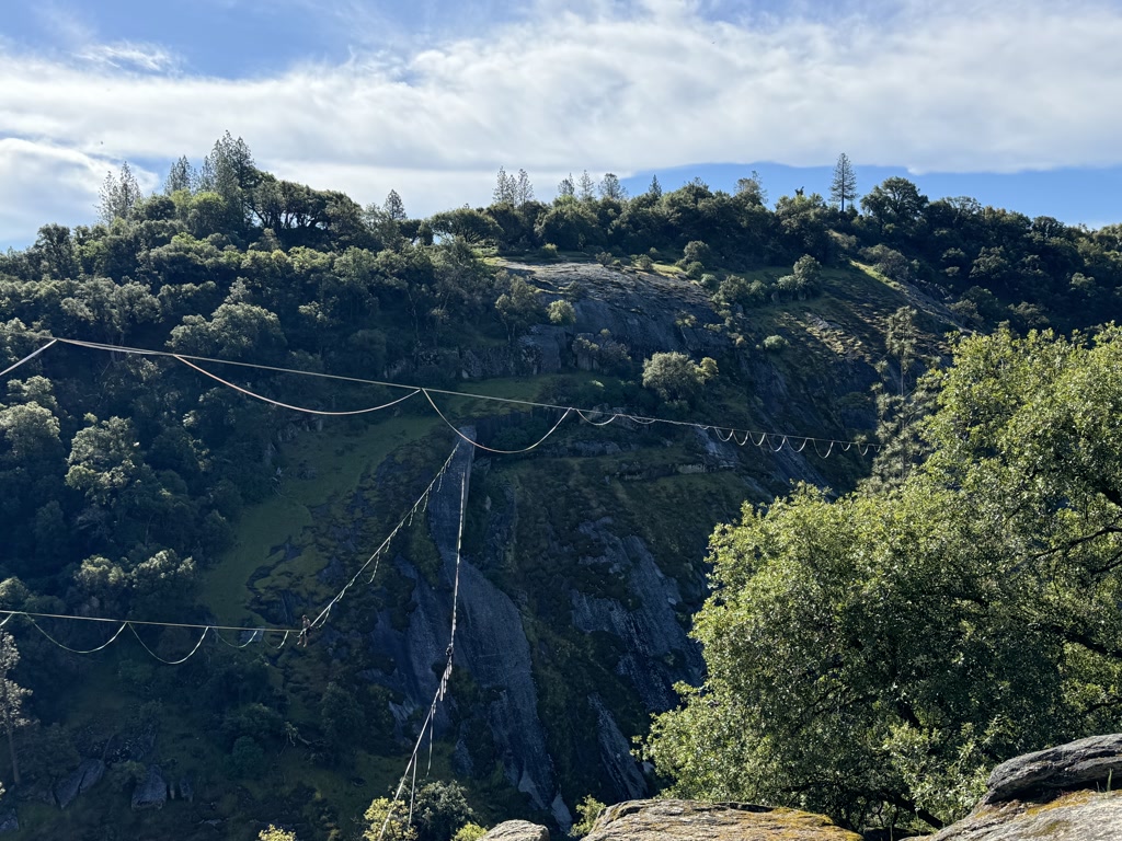 Three large, parallel slacklines are stretched across a wide, green valley, surrounded by rocky hillsides with scattered vegetation. The slacklines create a prominent feature against the natural landscape, connecting two opposing rock crests. A lush mix of shrubs and trees covers the slopes, and the sky above is partly cloudy with ample daylight.