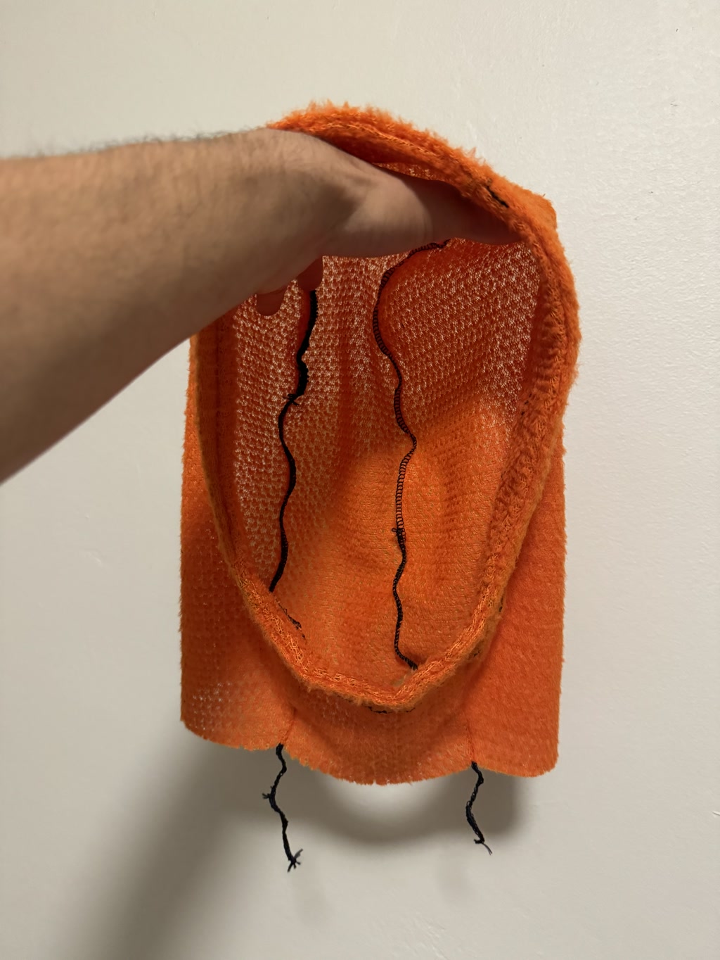 A work-in-progress orange sweatshirt hood is showcased, with someone's hand inside it to demonstrate shape. The textile fabric appears soft and porous, with characteristic fleece texture. Notable detailing includes a deliberately unfinished, frayed edge where the hood might be attached to a sweatshirt, exhibiting untrimmed threads hanging freely. Two parallel, wavy black stitch lines run down the middle, adding a touch of design to the otherwise solid-colored hood.