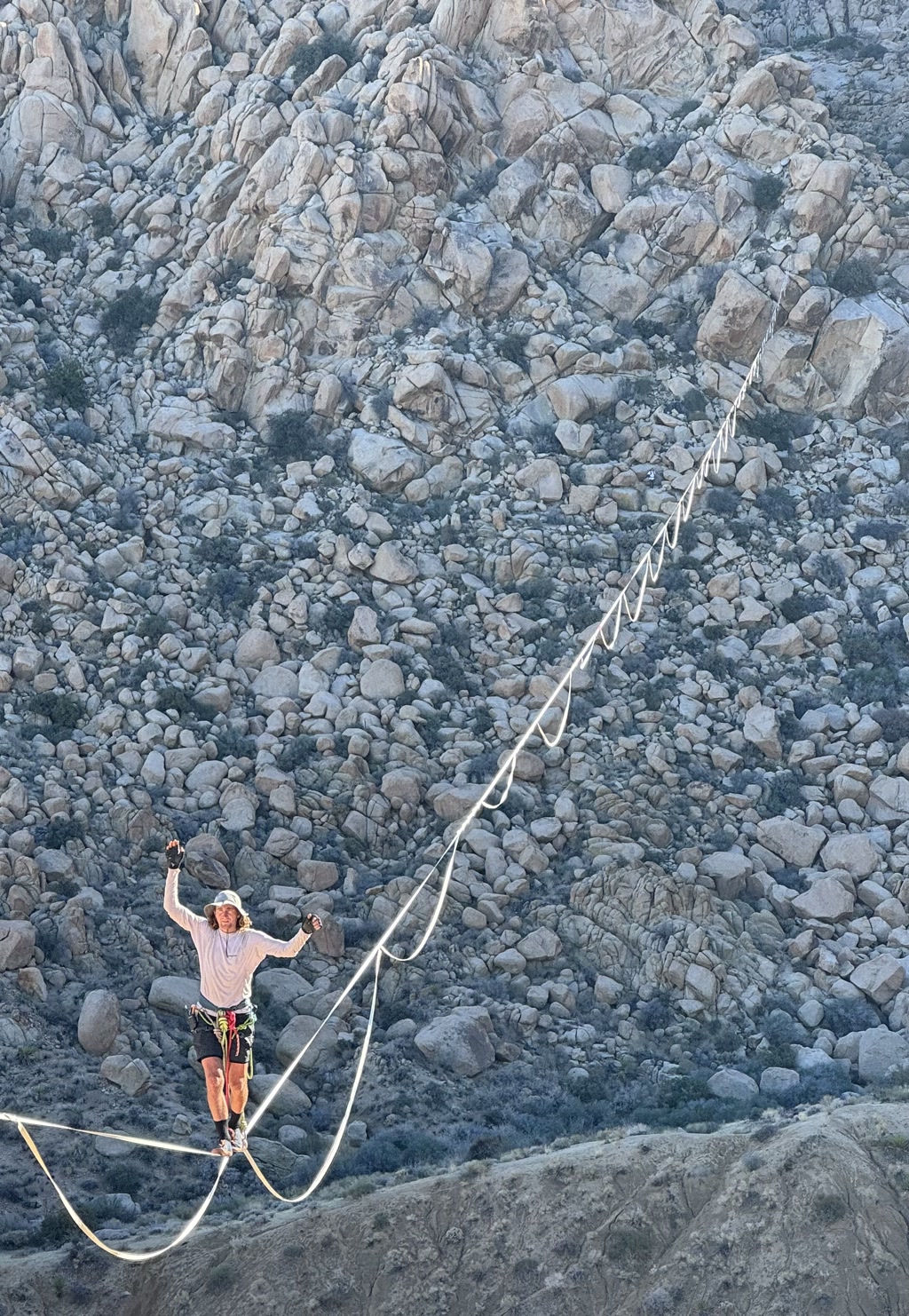 A person is engaging in highlining (slacklining at a significant height) across a large gap between rock formations. They are standing on a thin line stretched taut between the two points. Below, the terrain is rugged and strewn with numerous stones and boulders. The highliner is wearing casual athletic clothing, along with a safety harness that is presumably attached to the line for security. They are raising their arms, possibly in triumph or balance, set against the natural backdrop of the environment. The scene suggests a combination of adventure sports, focus, and the serenity of nature.