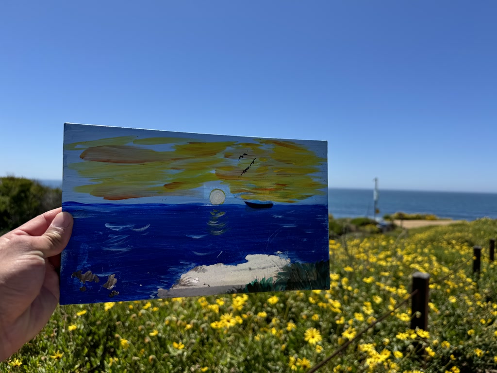 A hand is holding a small painting that depicts a serene coastal scene with vibrant blue water, a pale sandy beach, and a yellow sky, suggesting dusk or dawn. The painting is aligned with the actual coastal background, blending art with reality. The coastal background features a clear blue sky and a lively field of wild yellow flowers. There appears to be a cliff edge with a guarding rail, and beyond it the calm blue ocean extends to the horizon.