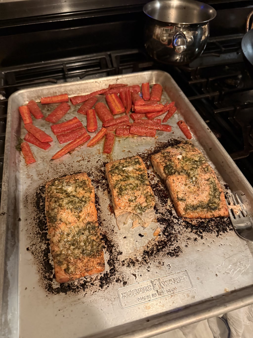 The image shows a baking tray with three pieces of baked salmon and numerous pieces of roasted carrots. The salmon appears to be seasoned with some herbs, and the carrots are cut into uniform strips. At the bottom of the tray, there is a label that reads 'NORDIC WARES' with a small logo and the text 'MADE IN THE USA.'